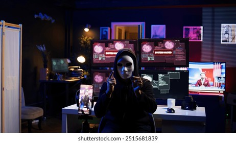 Evil programmer concealing identity using mobile phone to film ransom video in secret base. Scammer with anonymous mask using cellphone to record blackmail footage, threatening victims, camera B