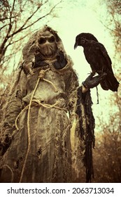 An evil Scarecrow with a canvas bag on his head and in a sackcloth robe stands in a dense forest with a black raven on his hand. Terrible forest sorcerer. Halloween Tales. Horror, thriller. - Shutterstock ID 2063713034