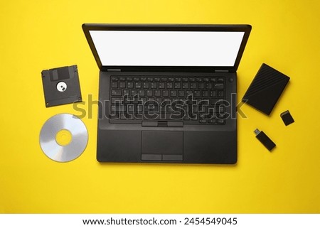Evolution of storage media. Laptop with CD disk, sd card, floppy disk, USB flash drive and external hard drive on yellow background. Flat lay