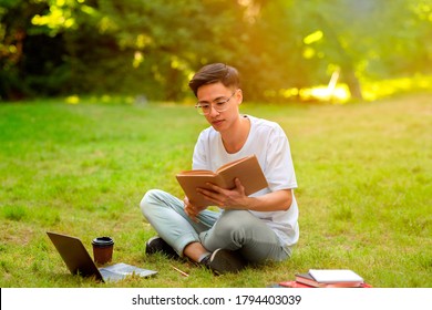 Exam Study. Chinese Student Guy Reading Book Outdoors In Park, Wearing Eyeglasses, Sitting On Lawn With Laptop And Coffee Next To Him