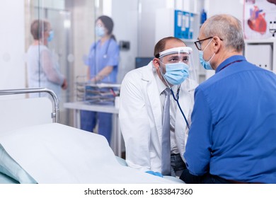 Examination Of Patient Heart Beat Using Stethoscope In Hospital Room And Wearing Face Mask As Safety Precaution Against Coronavirus. Medical Examination For Infections, Disease And Diagnosis.