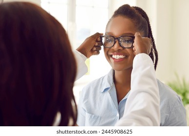 Examining eyeglasses. Happy African American female patient trying on glasses as recommended by ophthalmologist. Doctor puts on black-rimmed vision glasses on young woman. Ophthalmology concept. - Powered by Shutterstock