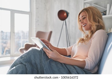 Examining her new tablet. Attractive young woman using digital tablet and smiling while sitting on the sofa at home - Powered by Shutterstock