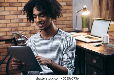 Examining his new tablet. Cheerful young African man holding digital tablet and looking at it with smile while sitting besidehis working place - Shutterstock ID 353560736