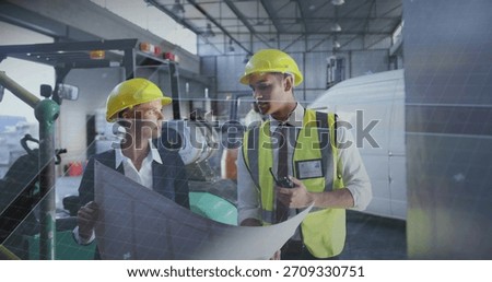 Examining two construction workers wearing yellow hardhats and vest in hangar, holding blueprint. Worksite, engineer, supervisor, plans, hi-vis, warehouse, machinery
