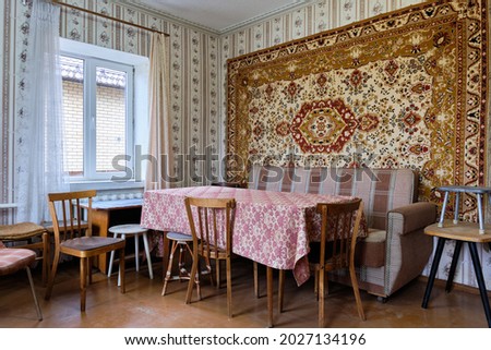 Example of Old Soviet Russian poor interior in Khruschev House. Aged table, chairs, sofa. Shabby floor. Tattered wallpaper and carpet as decor on the wall. Apartment of pensioners.