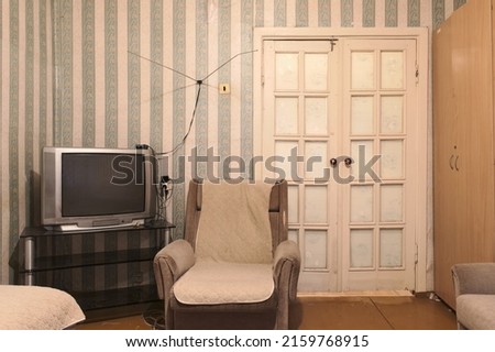 Example of Old Soviet Russian poor interior in Khruschev House. Aged TV, chairs, sofa. Shabby floor. Tattered striped wallpaper and antenna on the wall. Apartment of pensioners.