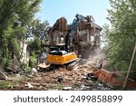 Excavator demolishing house. Large yellow bulldozer on top of building rubbish tearing down roof of home while water is sprayed to reduce dust. Active demolition background. Selective focus.