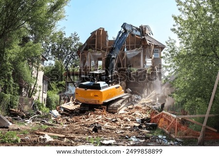 Excavator demolishing house. Large yellow bulldozer on top of building rubbish tearing down roof of home while water is sprayed to reduce dust. Active demolition background. Selective focus.