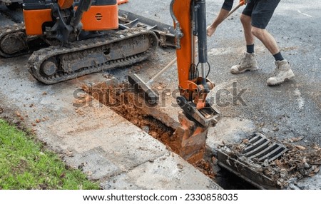 Excavator digging up dirt between cut concrete pavement. Man sweeping dirt with broom. Roadworks in Auckland.  
