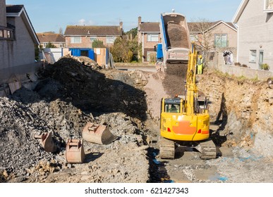 Excavator Is Digging A House Foundation In A Residential Area While A Dumper Truck Is Unloading Construction Gravel, Sand And Crushed Stones On The Construction Site.