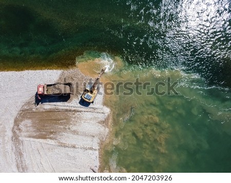 Excavator digging and loading sand into dump truck at river bank. Heavy machinery working at sand quarry. Aerial drone view of digger and truck loading gravel in nature.Environment and water pollution
