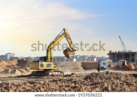 Excavator digging sand and loading into dump truck on construction site. Backhoe digs the ground, foundation for the construction of a new residential building. Tower cranes in action
