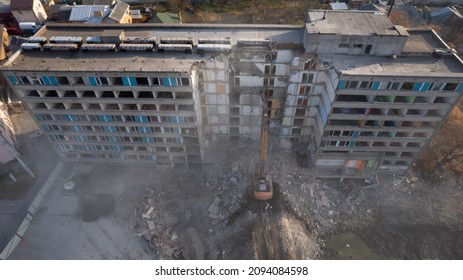 Excavator for dismantling buildings, using a special claw, dismantles a multi-storey building. The sun's rays break through a thick cloud of dust in front of the ruined building