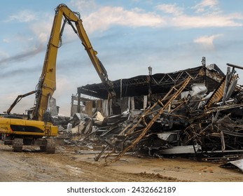 Excavator dismantling the charred remains of a burnt warehouse, twisted metal structures visible.