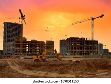 Excavator during earthmoving work at construction site on sunset background. Backhoe digging ground for foundation pit. Tower cranes in action during construction of a residential building