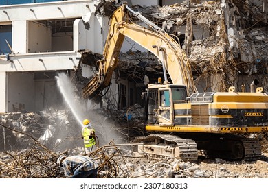 excavator equipped with a hydraulic hammer demolishes old building while a worker sprays water to minimize dust - Powered by Shutterstock