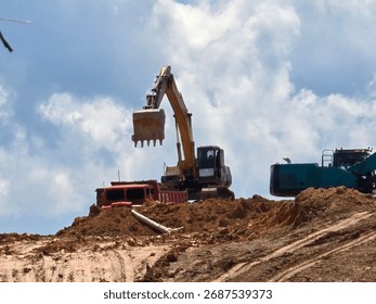 Excavator loading soil into a dump truck at a construction or mining site under a bright blue sky. The heavy machinery and rugged terrain represent industrial work, earthmoving, and land development c