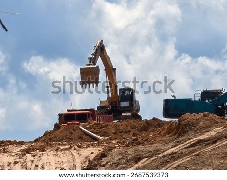 Excavator loading soil into a dump truck at a construction or mining site under a bright blue sky. The heavy machinery and rugged terrain represent industrial work, earthmoving, and land development c