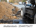 An excavator next to a deep long earthen trench for laying a large diameter water pipe. construction work to replace a city water pipe