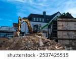 An excavator operates at a construction site, clearing debris from a demolished structure while a green crane is positioned nearby under a blue sky.