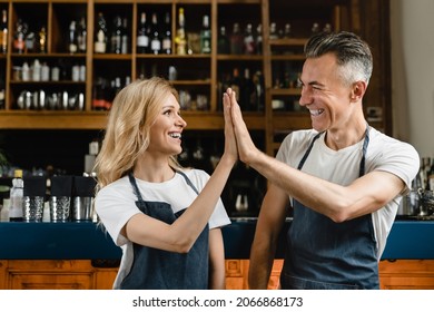 Excellent great job! Well done! Smiling mature caucasian small business owners waiters bartenders in blue aprons giving high five with arms crossed at the bar counter in restaurant.