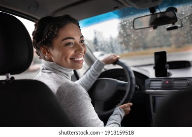 Excellent trip, auto travel and enjoy journey in town. Happy young african american woman driver turns to passenger in back seat. View of city road, window with raindrops and smartphone, copy space