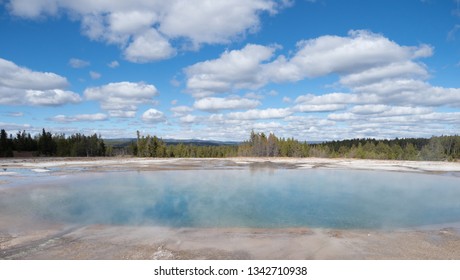 The Excelsior Geyser Crater In Yellowstone National Park, The Largest Geyser In The World, With Cumulus Clouds Overhead And Evergreen Trees In The Background.