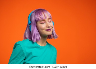 Exceptional woman with dyed very peri hair listening music in headphones and singing on orange background. Unique hipster girl dancing