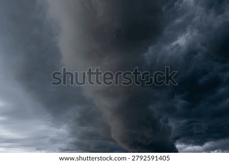 Exceptionally dramatic and cinematic storm sky image capturing the fearsome structure of a severe weather system at its most intense and visually arresting. A towering, near-black wall of storm cloud dominates the right side of the frame, its dense, vertical mass suggesting the powerful updrafts and extreme atmospheric energy associated with supercell thunderstorms or severe squall lines. The cloud formation tapers and swirls with a vortex-like quality in the lower frame, hinting at the rotational energy within the storm system and evoking the precursor formations of a tornado or funnel cloud. To the left, lighter grey clouds provide tonal contrast and a sense of the vast scale of the weather event, their turbulent, wind-driven forms adding further movement and unease to the composition. The near-absence of any warm tones gives the entire image a cold, stark and menacing quality that is both scientifically compelling and visually powerful. Shot with no horizon or ground reference, the image places the viewer entirely within the storm, amplifying its psychological impact. An outstanding image for meteorology and extreme weather content, news and editorial use, environmental and climate change campaigns, dramatic background and wallpaper use, film, gaming and music creative projects, and any design work requiring a visceral, impactful and awe-inspiring natural force visual.