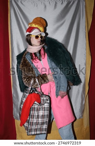 Excessive consumption of things. A young man with long hair and glasses wearing clothes of different colors and sizes against a gray background.
