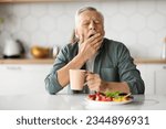 Excessive Daytime Sleepiness. Tired Senior Man Yawning At Table In Kitchen, Elderly Gentleman Feeling Sleepy While Eating Lunch At Home, Covering Mouth With Hand, Suffering Hypersomnia Disorder