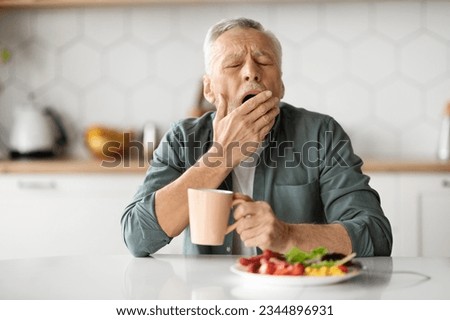Excessive Daytime Sleepiness. Tired Senior Man Yawning At Table In Kitchen, Elderly Gentleman Feeling Sleepy While Eating Lunch At Home, Covering Mouth With Hand, Suffering Hypersomnia Disorder