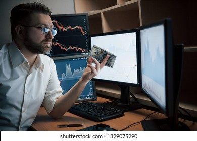 From exchange to real cash. Bearded man in white shirt and glasses holds money in the office with multiple computer screens in index charts.