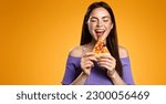 Excited 25 years woman bites tasty slice of pizza, orders delivery takeout from restaurant, hungry girl eats isolated on orange background.