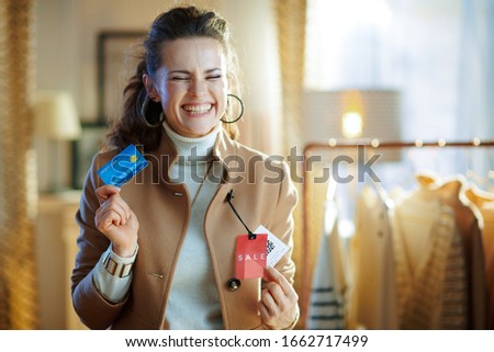 excited 40 years old woman in white sweater and skirt trying beige coat with red sale price tag and holding blue credit card in modern fashion store.