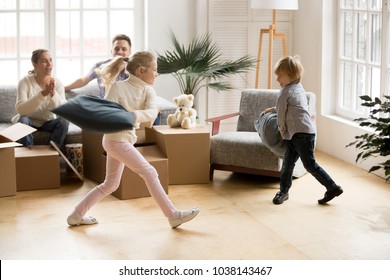 Excited Active Children Having Pillow Fight On Moving Day, Happy Girl And Boy Playing Together In New Home While Parents Laughing Supporting Kids, Family Packing Boxes Or Unpacking After Relocation