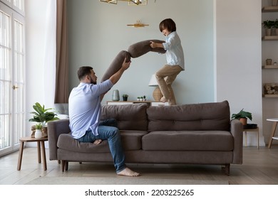 Excited Active Son Kid And Dad Fighting With Pillows On Sofa, Enjoying Funny Battle With Cushions. Father And Child Playing Games At Home, Having Fun Together, Fatherhood, Family Concept. Full Length