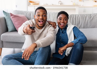 Excited African American Black Couple Watching TV Show Or Movie, Holding Remote Contoller, Sitting On The Floor At Home And Leaning On Sofa In Living Room, Amazed Guy And Lady Looking At Camera