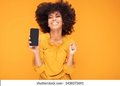 Excited African American Girl Hold Mobile Phone With Blank Empty Screen Isolated On Yellow Studio Background . Emotions, Lifestyle Concept. Copy Space.