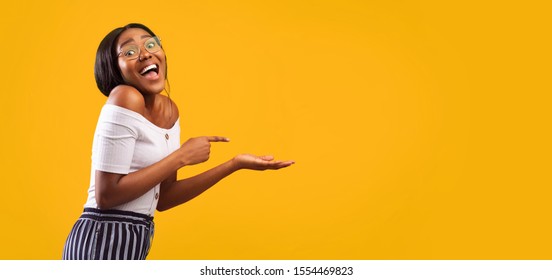 Excited African American Girl Pointing Finger At Open Palm Standing Over Yellow Background. Studio Shot, Panorama With Empty Space