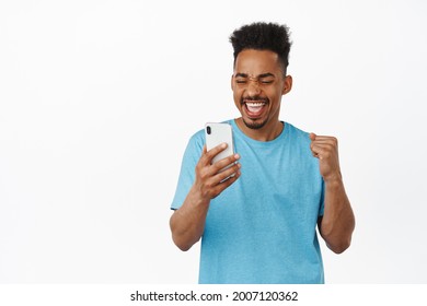 Excited African American Guy Fist Pump, Winning Money Online, Rejoicing With Smartphone In Hand, Say Yes With Rejoice And Looking At Mobile Phone App Screen, White Background