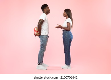 Excited African American guy holding wrapped gift box, hiding present for his girlfriend behind back, loving couple standing isolated over pink studio background, full body length, profile side view