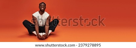 excited african American guy in trendy casual clothes sitting on red and orange backdrop, banner