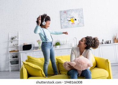 Excited African American Kid In Headphones Holding Smartphone Near Laughing Mother