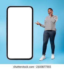 Excited African American Lady Standing Near Huge Smartphone Pointing Finger At Empty Screen And Touching Face On Blue Background. Female Advertising Mobile App. Square Shot, Full Length, Mockup