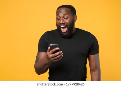 Excited African American Man Football Fan In Casual Black T-shirt Isolated On Yellow Background Studio Portrait. People Lifestyle Concept. Mock Up Copy Space. Using Mobile Phone Typing Sms Message