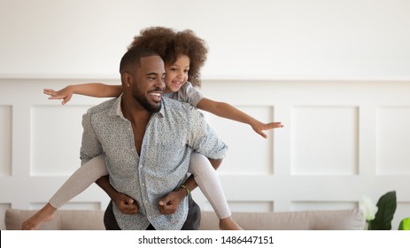Excited african american man giving piggyback ride to happy cute small daughter, enjoying active leisure time. Laughing black father carrying on back little baby girl playing, having fun together.
