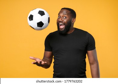 Excited African American Man Guy Football Fan In Casual Black T-shirt Isolated On Yellow Background. Sport Family Leisure Lifestyle Concept. Cheer Up Support Favorite Team Throwing Up Soccer Ball