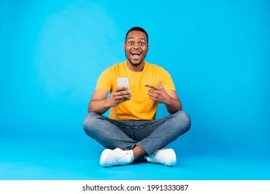 Excited African American Man Holding Smartphone, Pointing Finger At His Mobile Phone In Excitement Sitting Over Blue Background In Studio. Joyful Guy Recommending Application For Your Cellphone